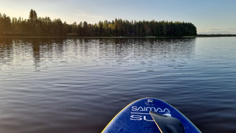 kesäilta järvellä, kuva otettu sup-laudalta. rantapuiden välistä häämöttää mökki.